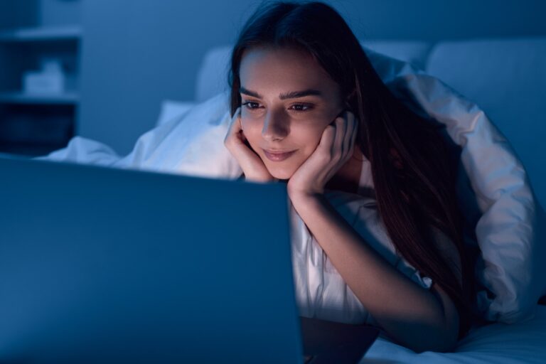 Young Hispanic lady watching video on laptop while resting on bed